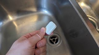 A dishwasher tablet held over a sink drain