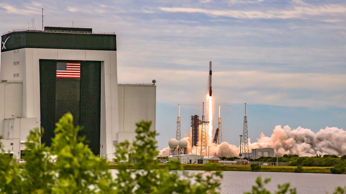 A SpaceX Falcon 9 rocket launches into space with a hangar and trees in frame