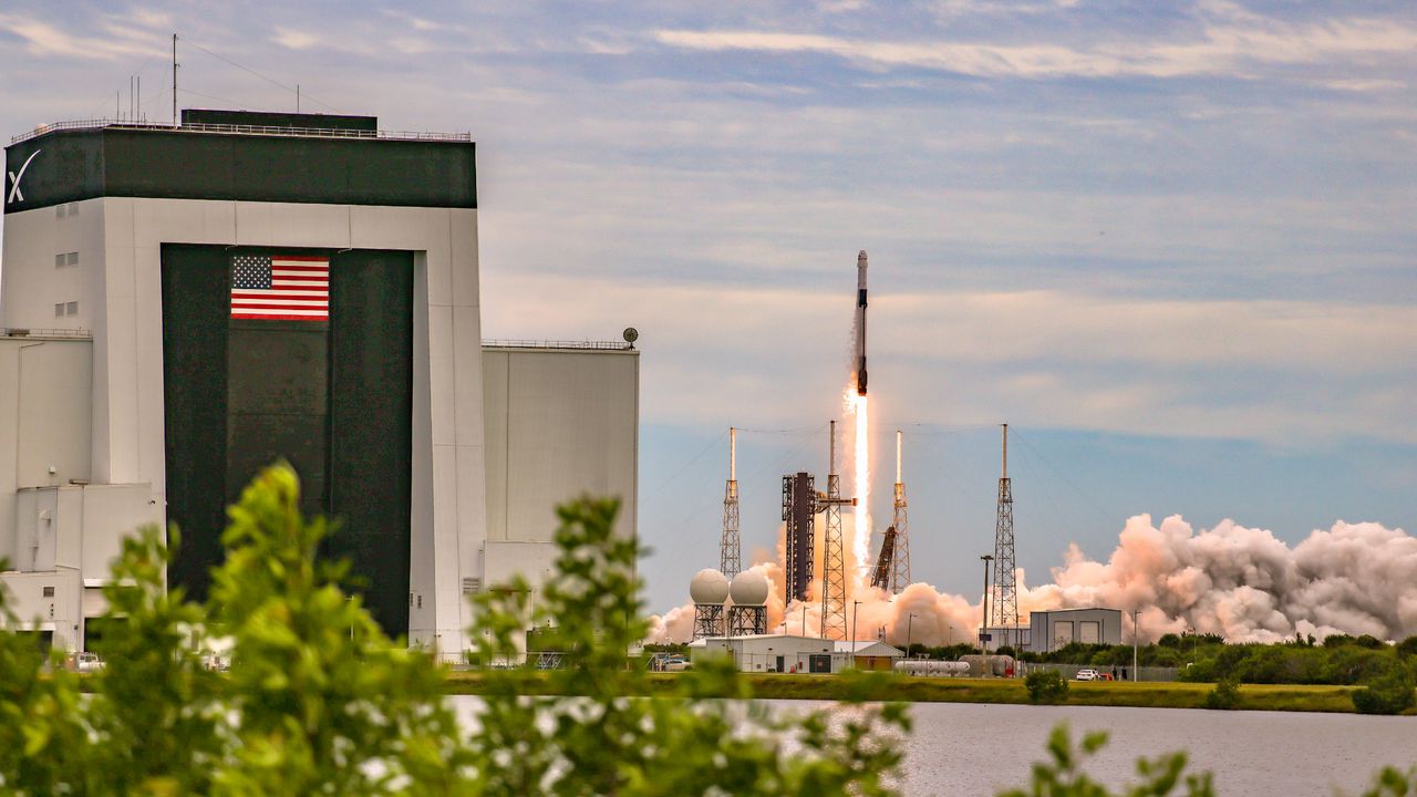 A SpaceX Falcon 9 rocket launches into space with a hangar and trees in frame