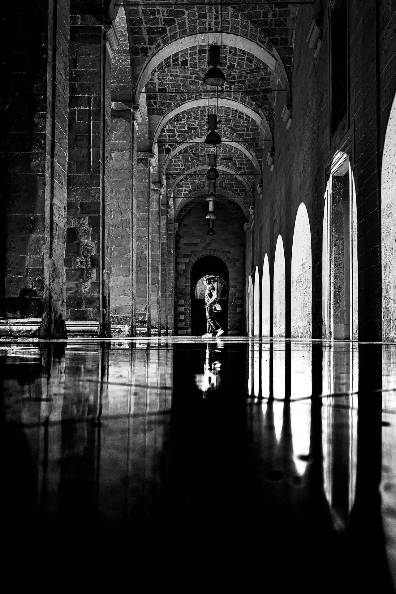 Person walking through a stone arch hallway with reflections on the shiny floor, in black and white