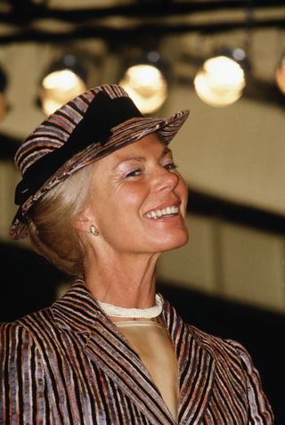 THe Duchess of Kent in a brown and black striped suit and matching hat with a black bow smiling and looking up