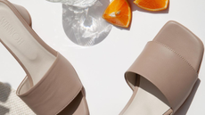A pair of beige leather mules on a white background with a glass of water and sliced oranges