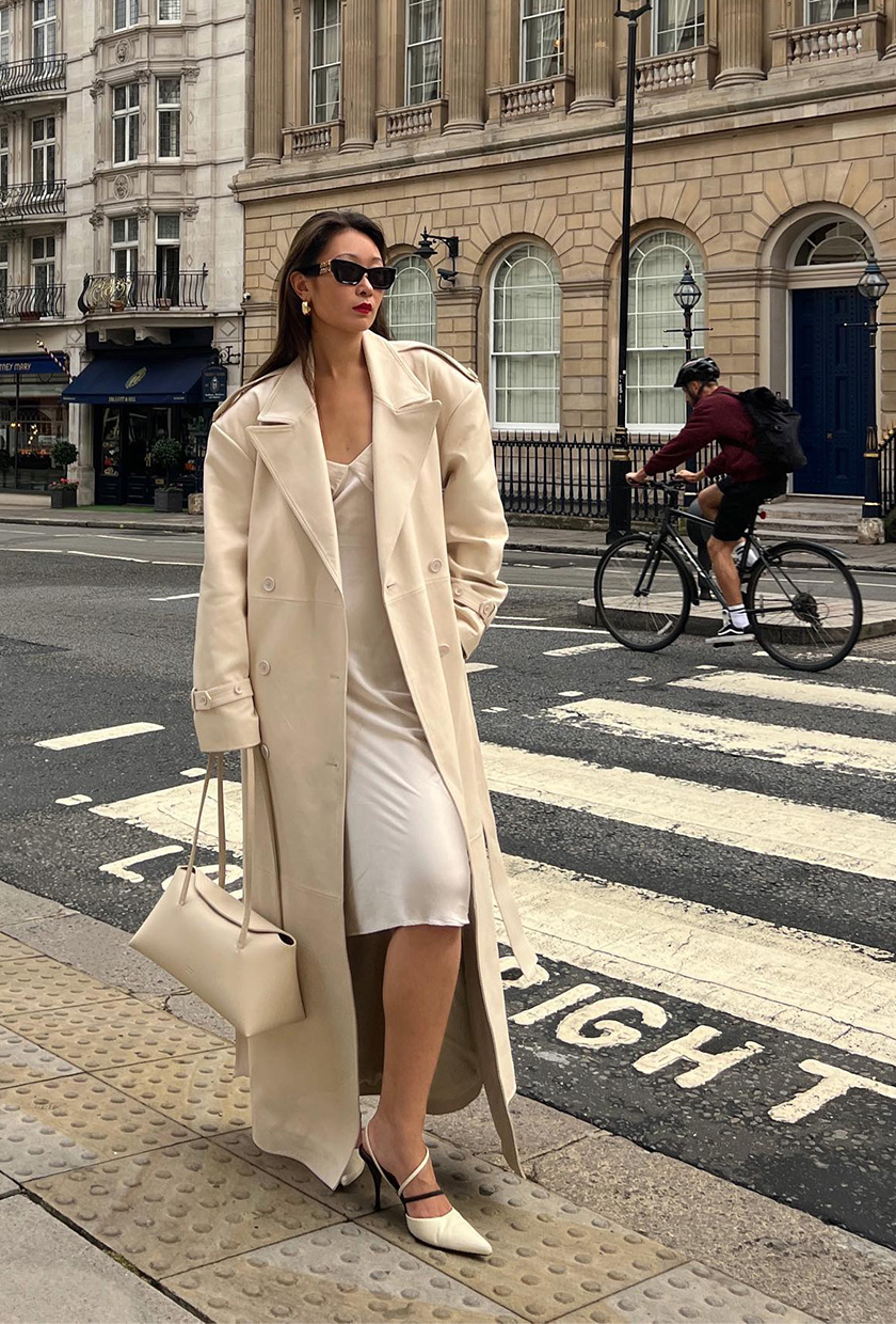 A woman's white dress outfit idea with a white slip layered under a cream trench coat with a matching bag and pointed pumps.