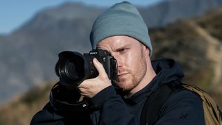 A man holding the Sony Alpha 7R VI mirrorless camera up to his eye to take a photo. He is standing outdoors wearing cold weather clothing, with mountains behind him.