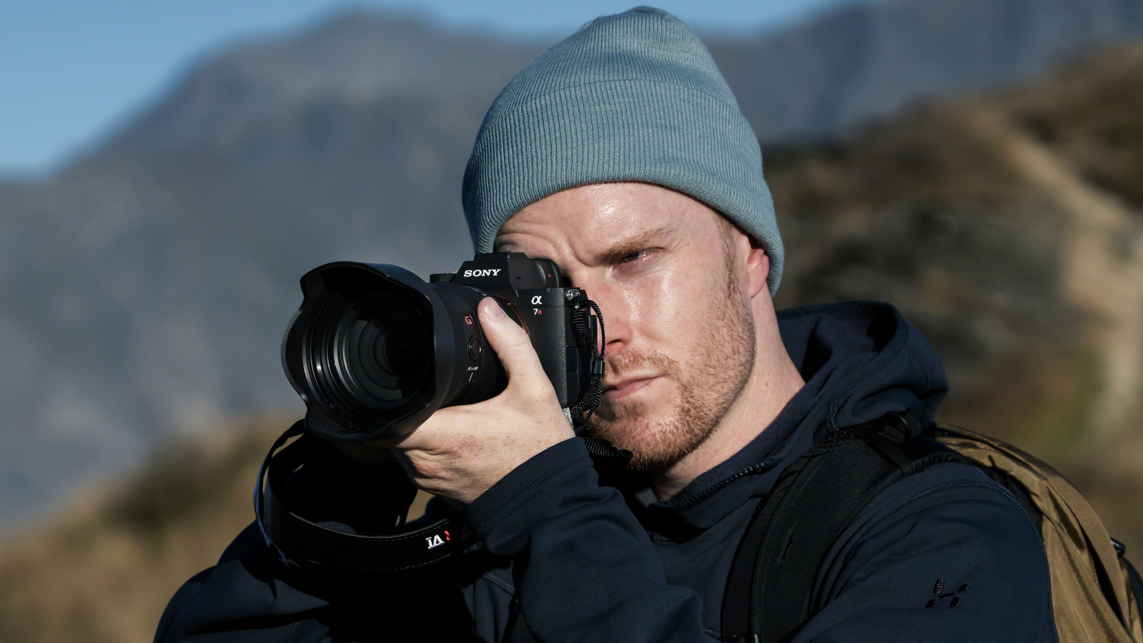 A man holding the Sony Alpha 7R VI mirrorless camera up to his eye to take a photo. He is standing outdoors wearing cold weather clothing, with mountains behind him.
