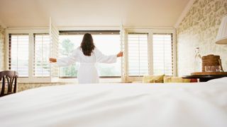 A brunette woman in a white fluffy bathrobe opens white shutters to let sunlight into her rustic European hotel room.