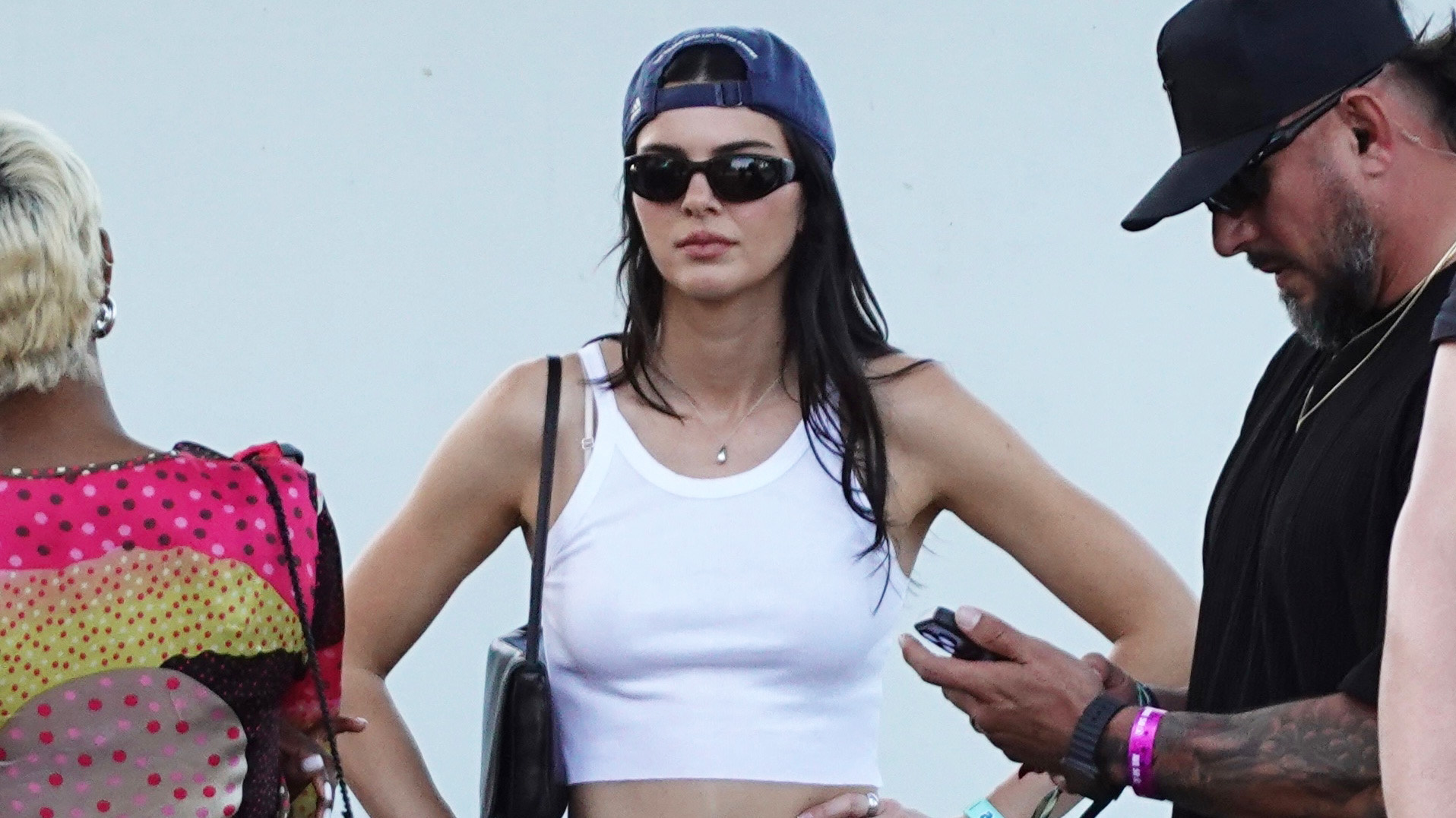 Kendall Jenner wearing a white tank top, baseball cap, and white cutoff denim shorts with black boots at Coachella weekend one 2026.