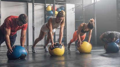 Four people in a gym perform planks and push-ups on medicine balls.
