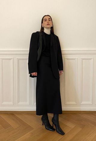 A cute work winter outfit is shown in a photo of a woman standing inside against a white wall wearing a white turtleneck sweater under a black crewneck sweater dress styled with a black blazer, black shoulder bag, black ankle boots, and silver jewelry