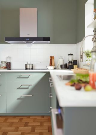 a sage green ikea kitchen with white worktops. There are drawers and cabinets in the space. Above the drawer is a stone and a silver extractor fan.