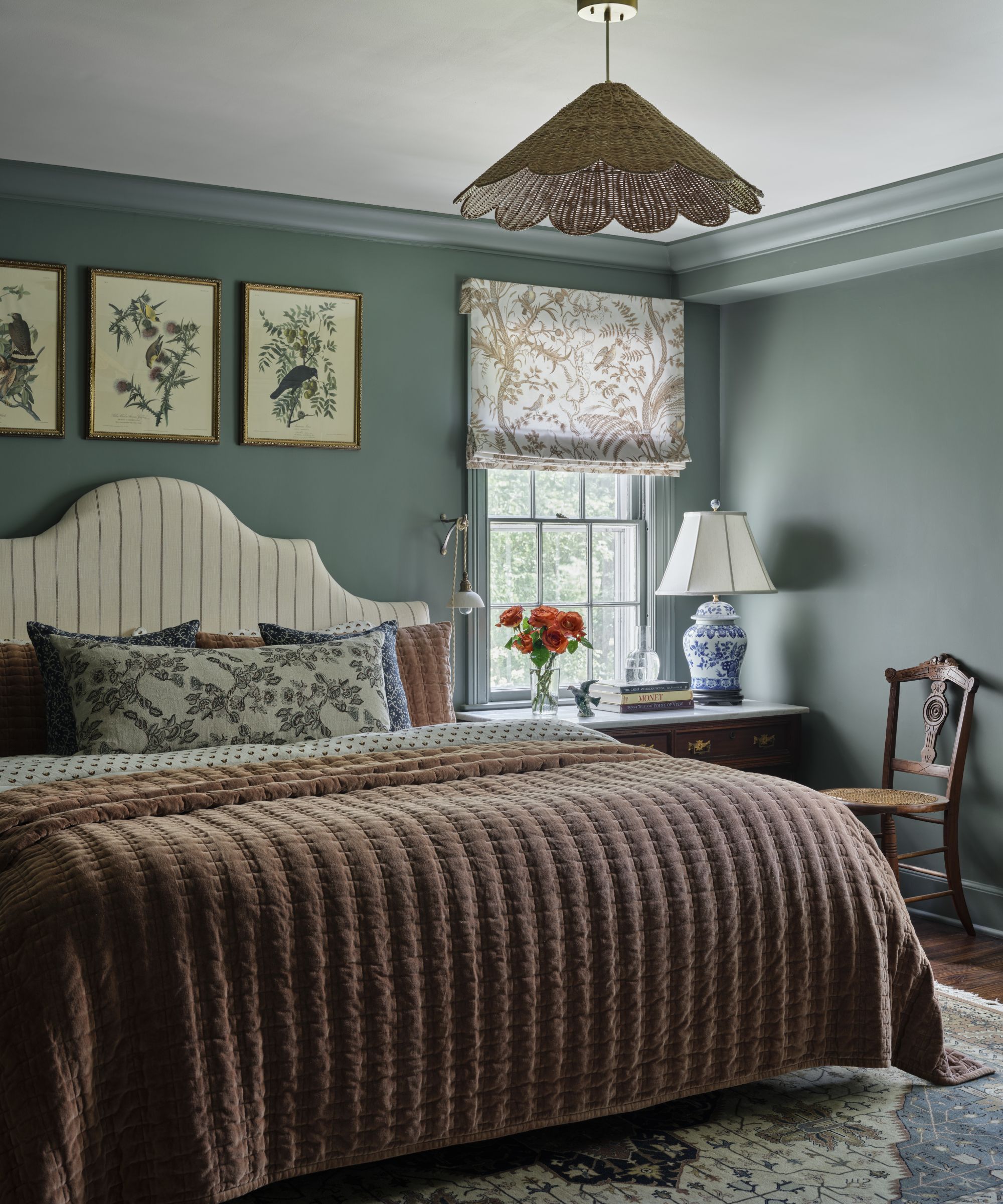 a warm blue green bedroom with rattan scalloped light, botanical wall prints and a striped headboard