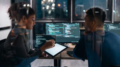 Male and female software developers discussing strategy in an open plan office space, with desktop computer showing source code on screen in background.