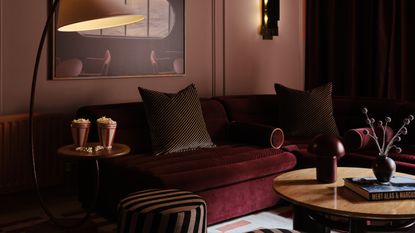 Image of a dusty pink painted media room with a large aubergine sofa. There is a painting on the wall above the sofa, a curved floor lamp beside the sofa, and a round wooden coffee table in front of the sofa.