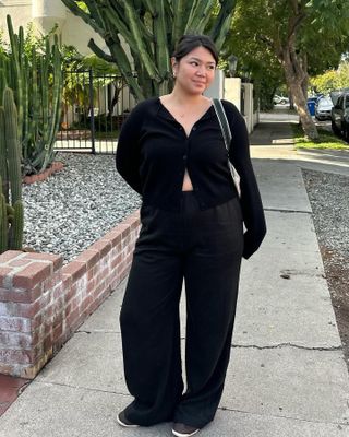 influencer Marina Torres poses on a sidewalk wearing a black cardigan sweater, black satin pants, and sneakers