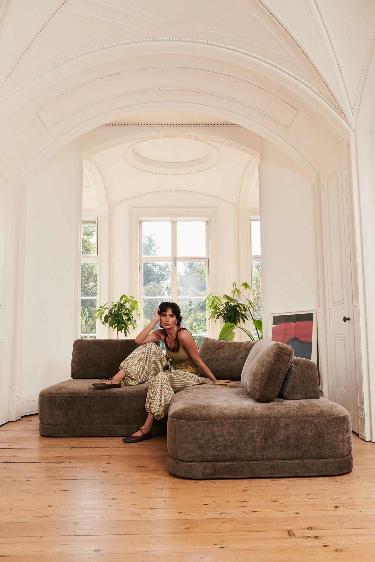 Image of a person in a green tank and green balloon pants sitting on a brown sectional sofa in a white living room with high ceilings and wooden floors