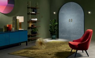 Living space, green walls, red chair, textured gold rug, blue cabinet, green potted plant, wall lights, wall art, arch grey doors, floor standing wooden shelving unit with gold frame, lamp with white shade