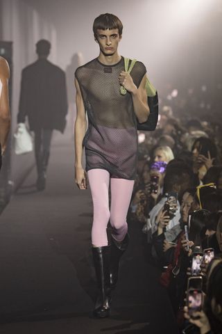 A cat walk focussing on a model wearing a net-like sleeveless top, pink leggings and knee-high black boots, carrying a satchel bag over the shoulder.