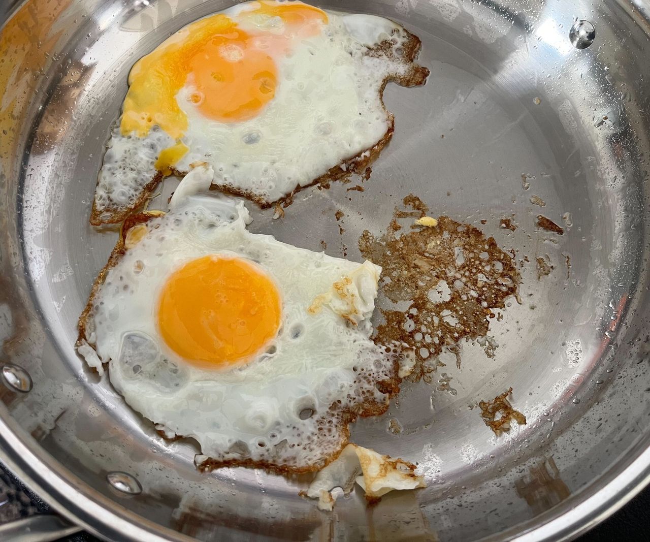 Two fried eggs in a stainless teel frying pan besides some brown stickadge.