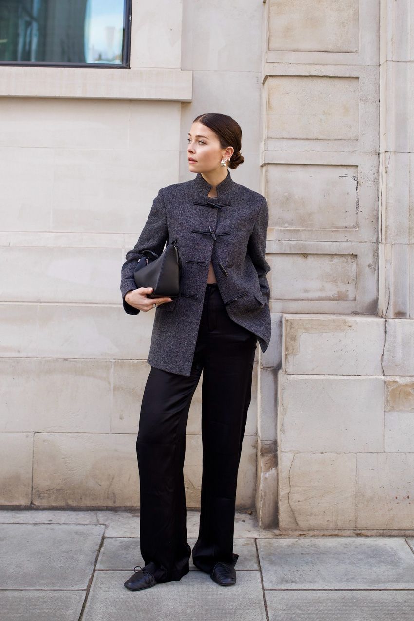 A street style photo of a woman standing outside wearing a gray tang jacket styled with black wide leg trousers, black ballet flats, a black shoulder bag, and silver earrings.