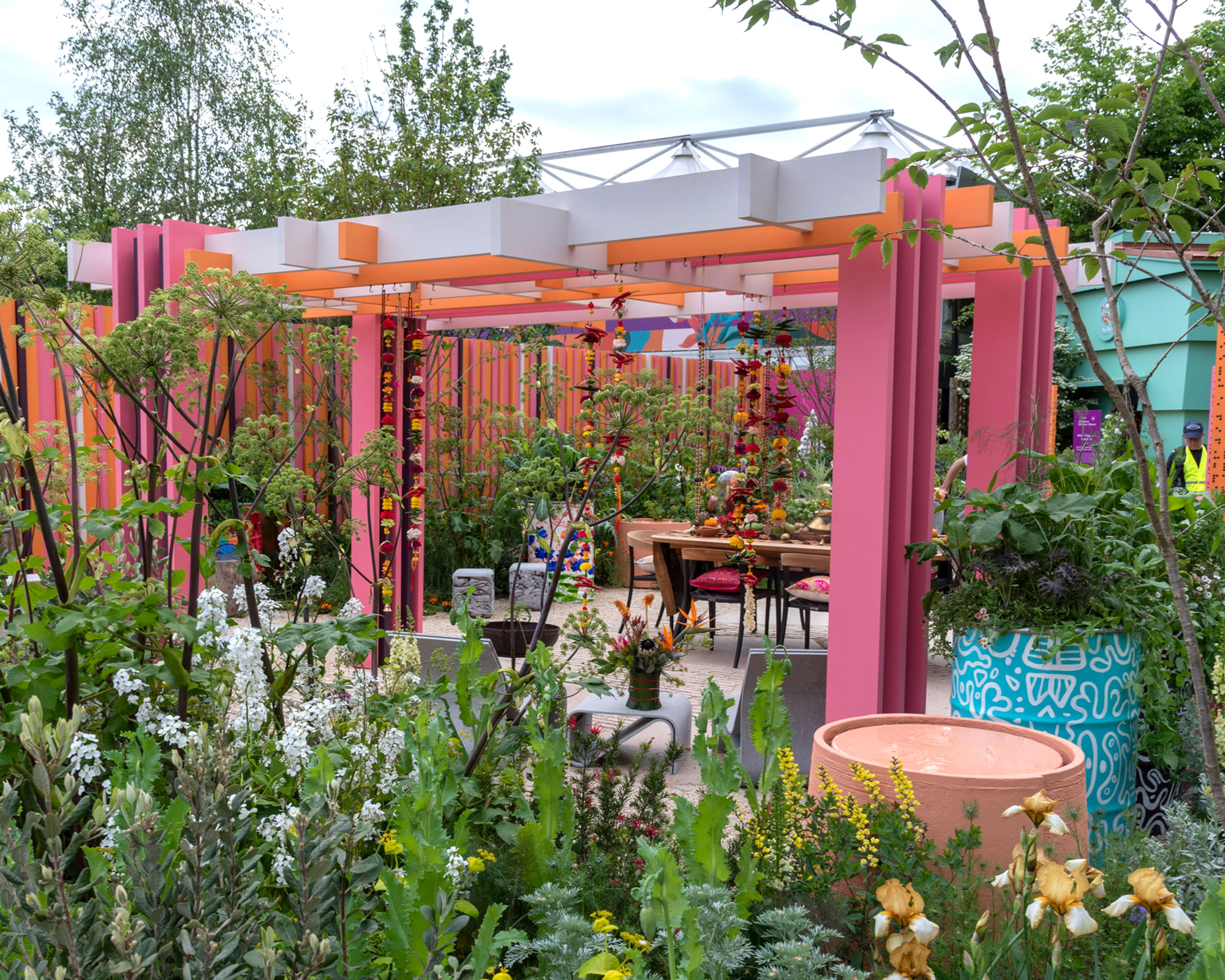 pink and orange painted pergola in garden filled with naturalistic planting, with garden furniture and painted fence