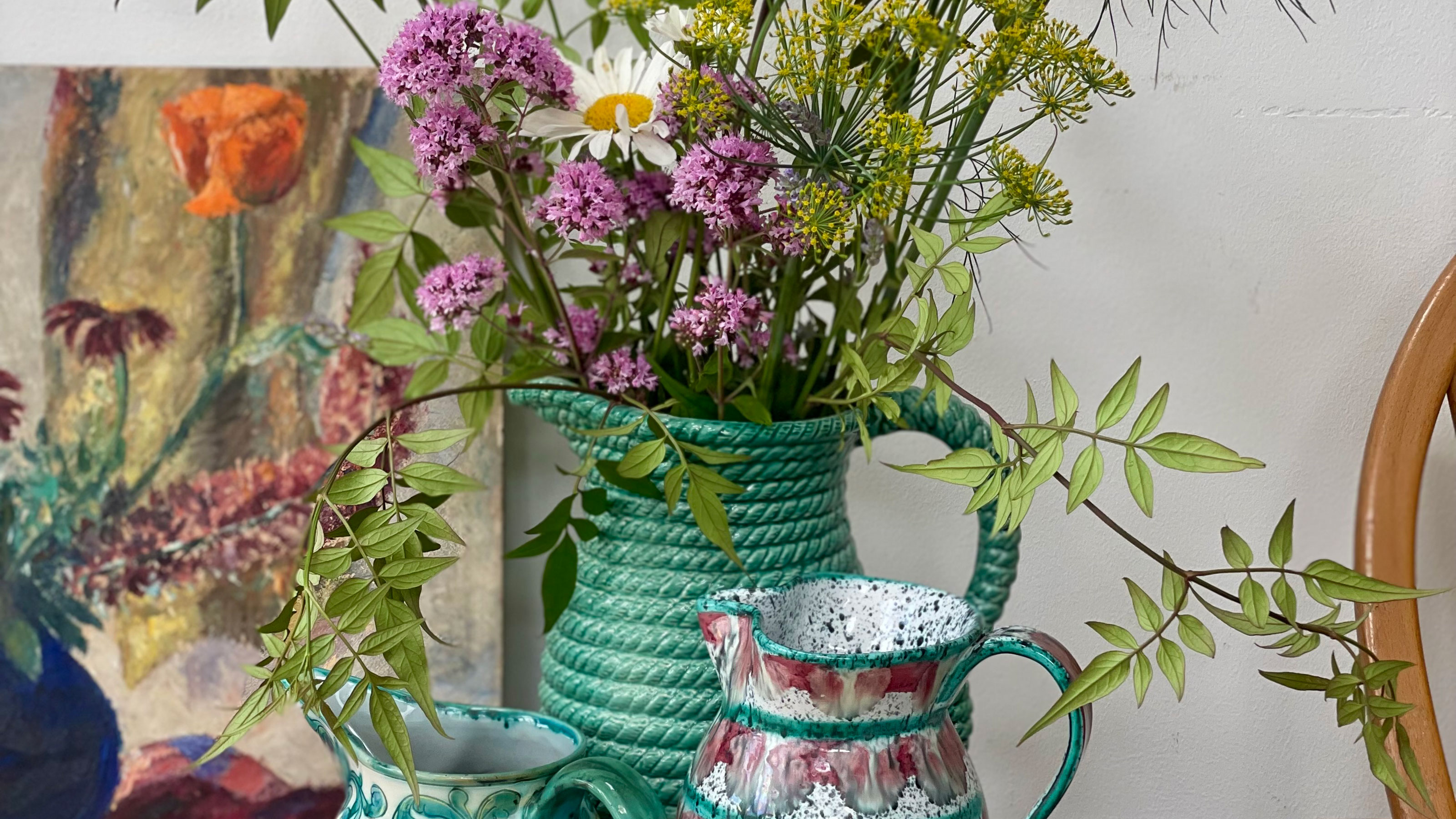 Three ceramic jugs, one with bunch of flowers, in front of piece of art