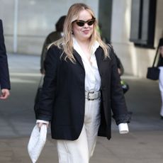 Nicola Coughlan walks down the street wearing white jeans with silver platform heels, a white shirt and a black blazer.