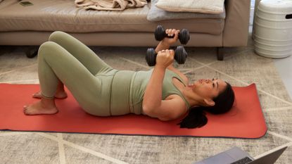 woman wearing a khaki leggings and top on an orange exercise mat holding two dumbbells above her chest. An edge of a sofa is behind her. 