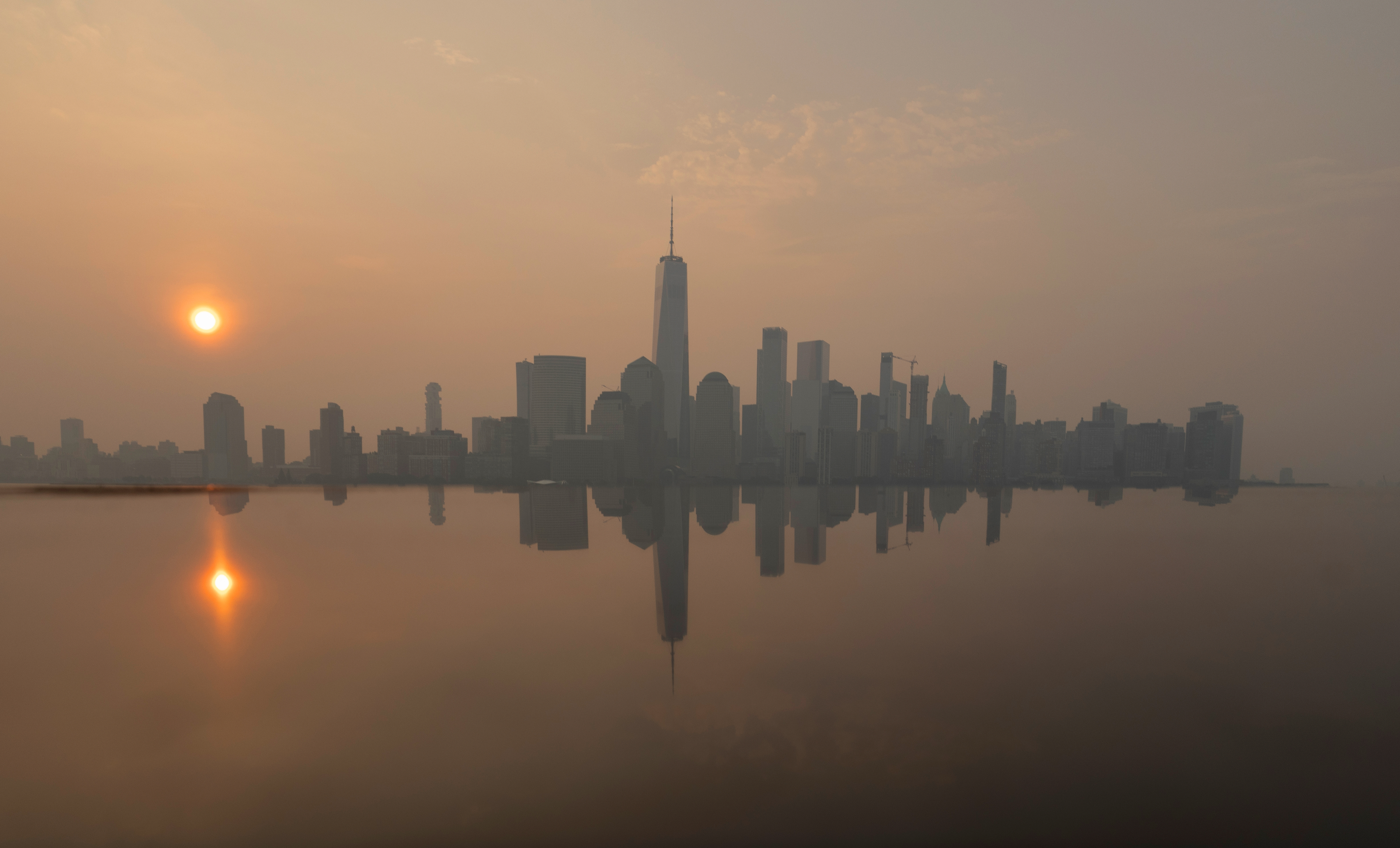 JERSEY CITY, NJ - JULY 18: The skyline of lower Manhattan and One World Trade Center are reflected on the top of a monument as wildfire smoke from Canada shrouds the sun as it rises behind in New York City on July 18, 2026, as seen from Jersey City, New Jersey. (Photo by Gary Hershorn/Getty Images)