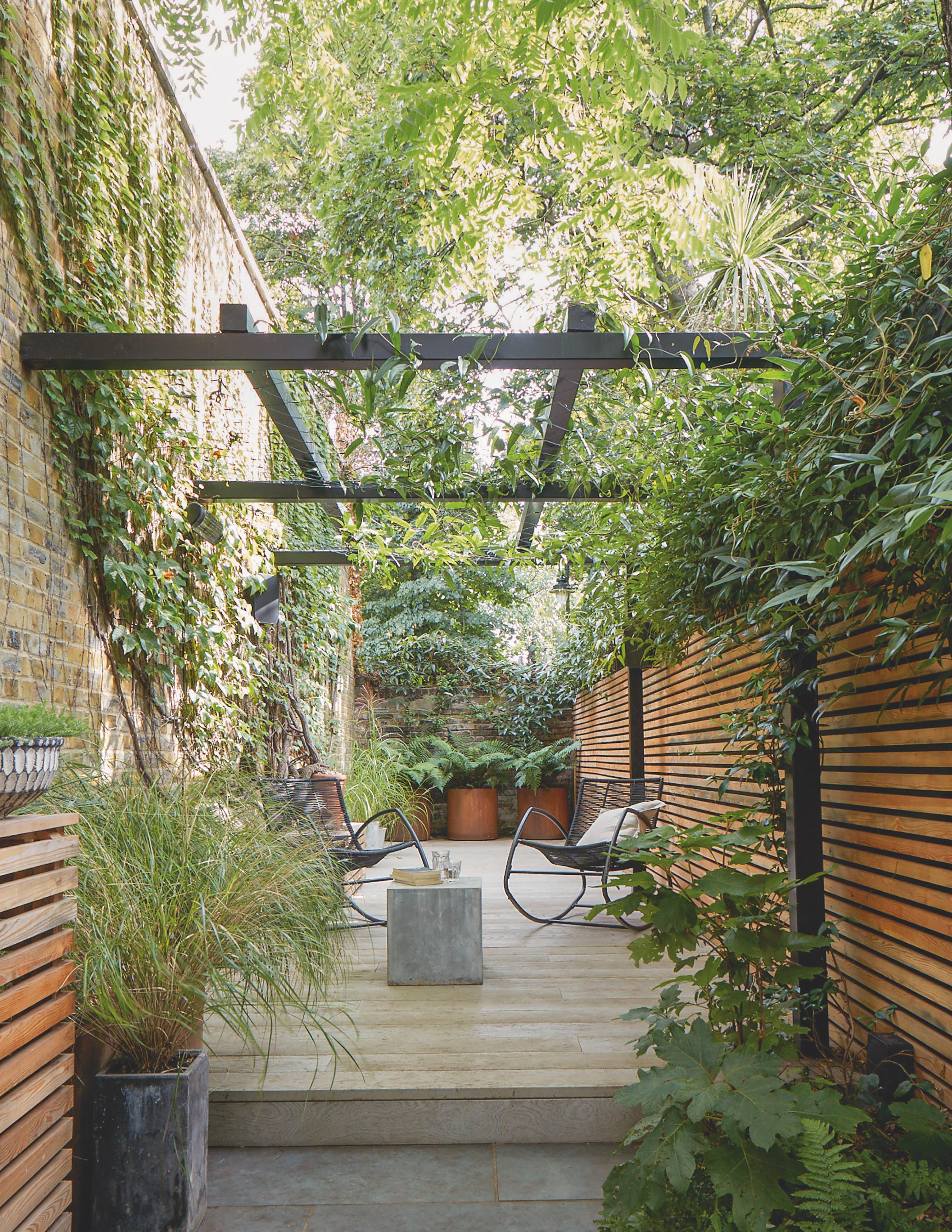 Raised, tiled seating area with black pergola and greenery above, and wooden fencing along the length of the outdoor space.
