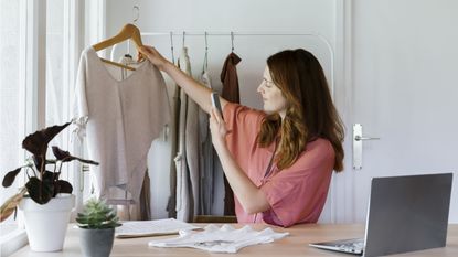 A woman at a desk holds up a blouse on a hanger and takes a photo of it.