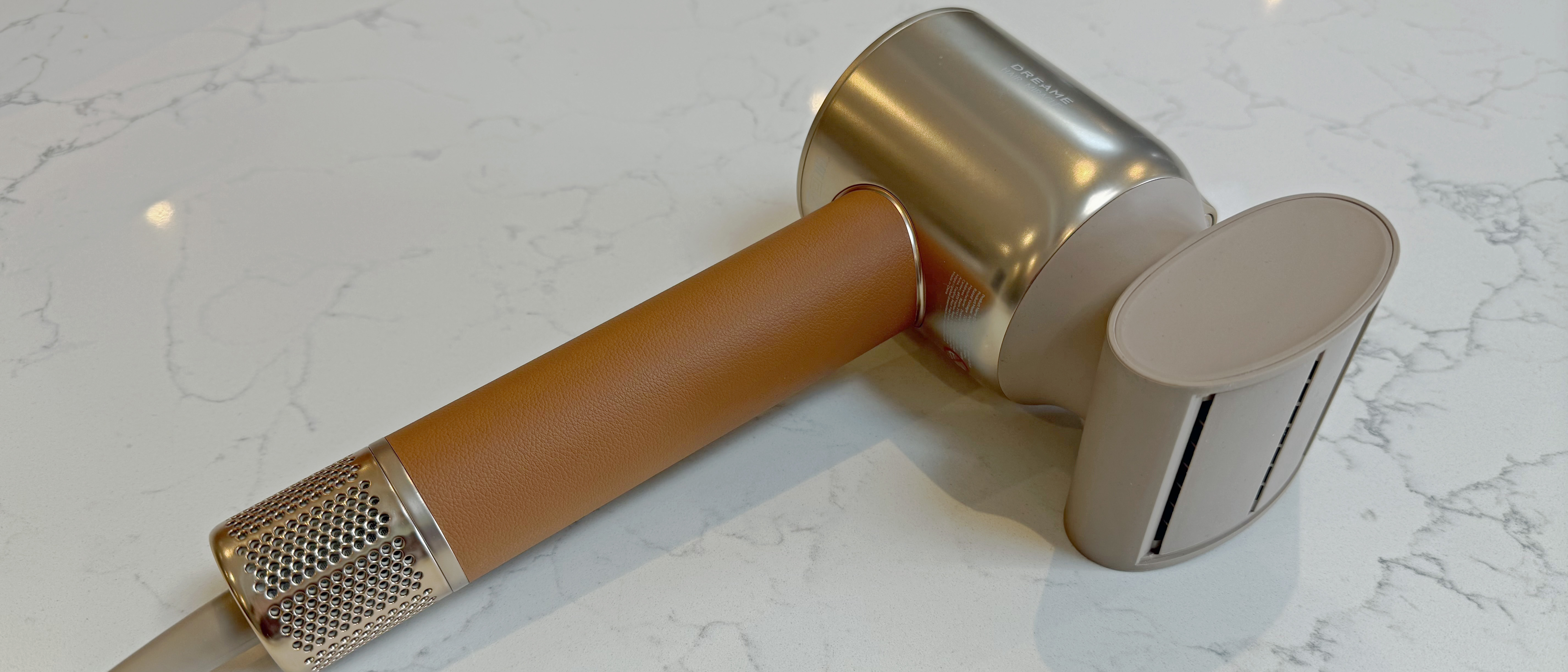 Dreame Miracle Pro hair dryer in gold and tan design lying on a quartz surface, showing the barrel and handle with the anti-frizz attachment 