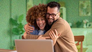 Young couple looking excited and hugging whilst sitting in front of a laptop.