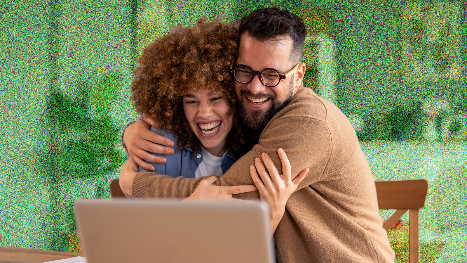 Young couple looking excited and hugging whilst sitting in front of a laptop.