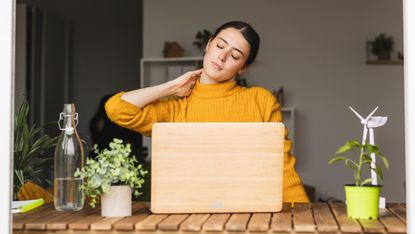 A woman sits at a wooden plank desk with a laptop open in front of her. She is stretching her neck towards her right shoulder and massaging it with her left hand. There are plants either side of the laptop and a glass bottle of water.