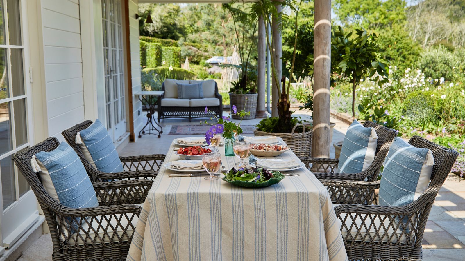 large nancy meyers style porch with woven wicker patio dining set styled with a beige and blue striped table cloth and blue cushions