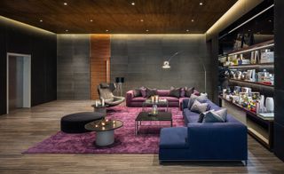 A lounge area with a blue sofa, a purple sofa, two square coffee tables, a round coffee table, a leather chair, a floor lamp, a purple rug and wall shelving.
