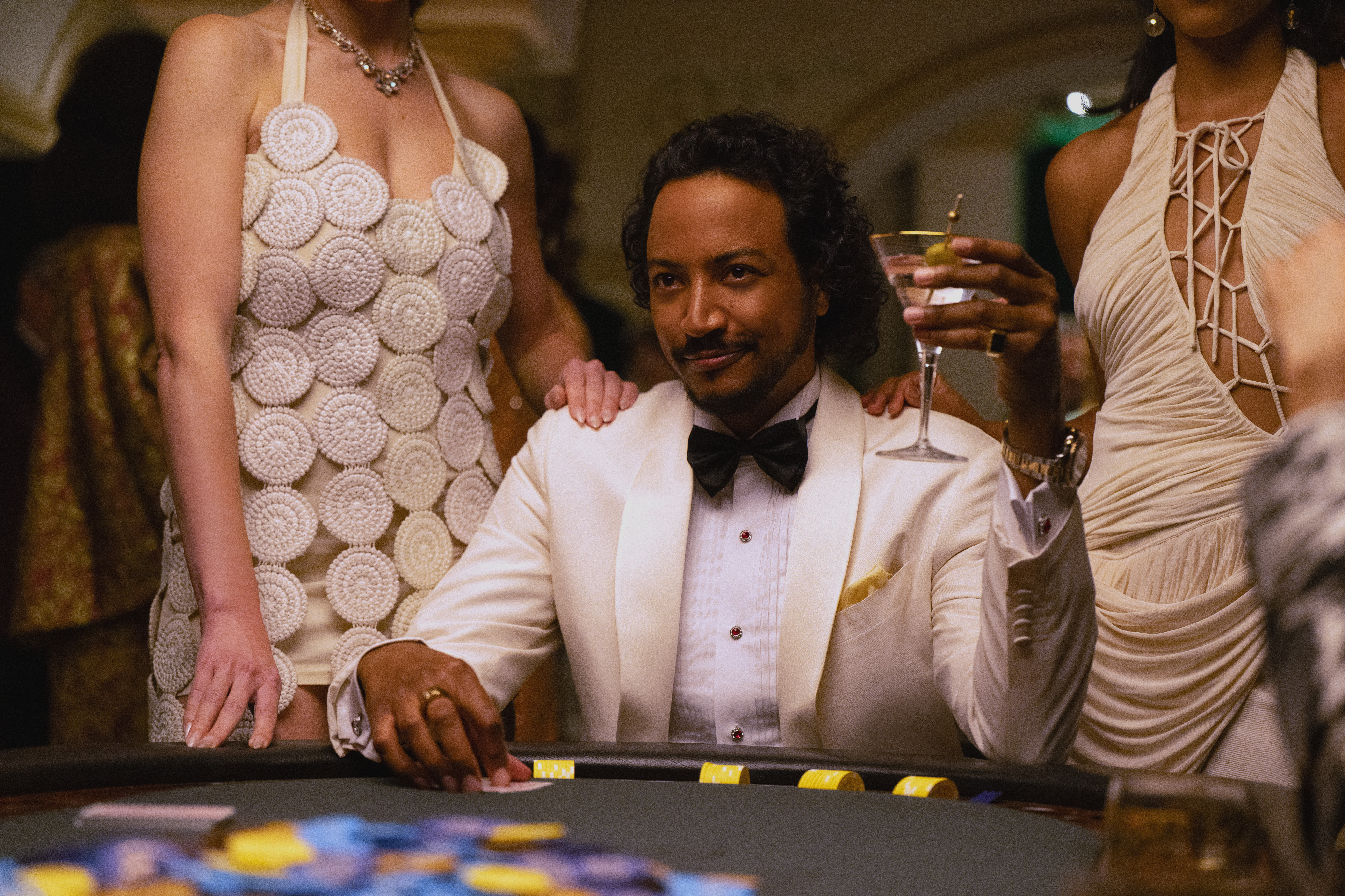A man wearing a white tuxedo (Samba Schutte as Diabat&amp;eacute;) sits at a poker table and raises a martini glass as women in white dresses stand around him, in a still from 'Pluribus.'