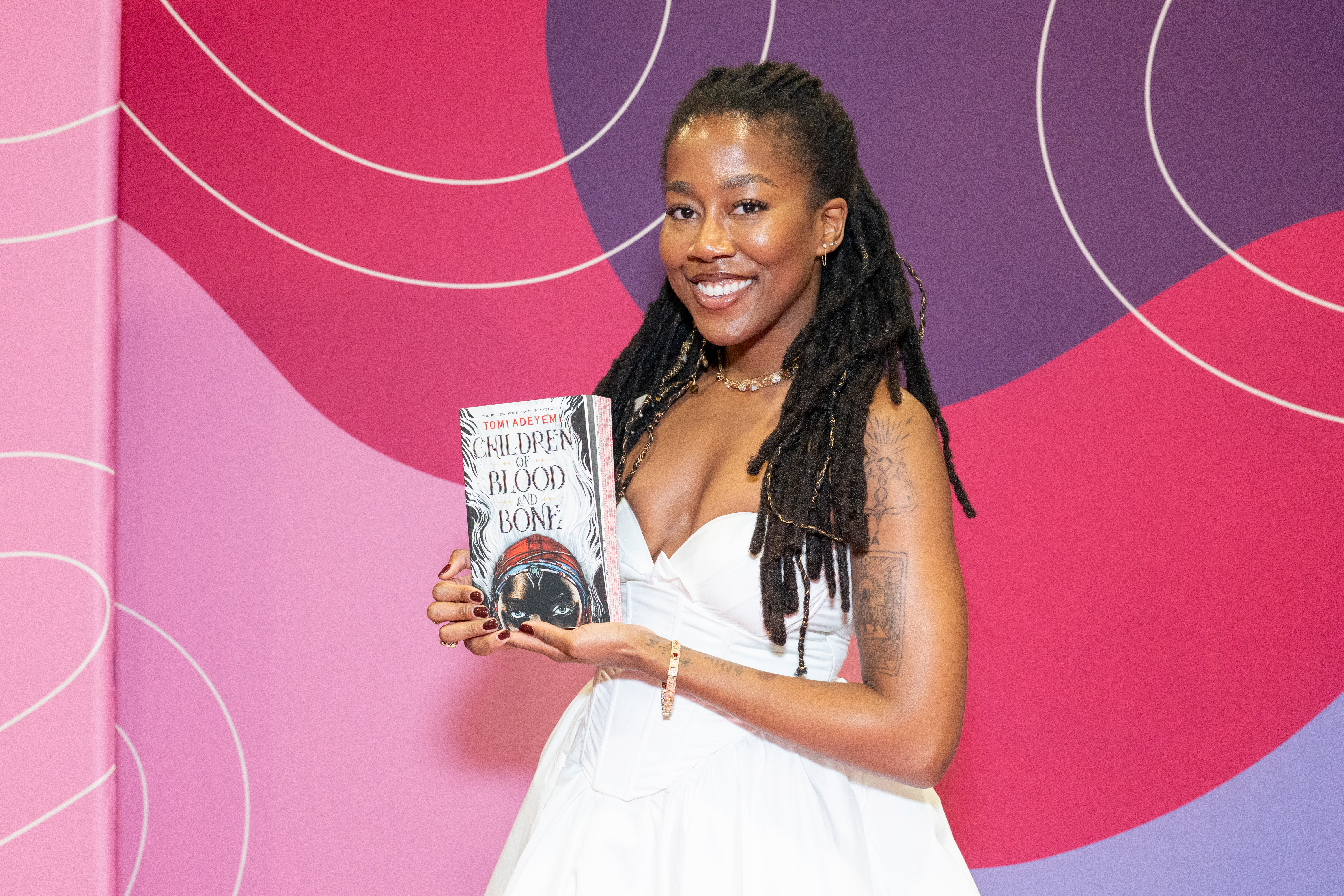 children of blood and bone author tomi adeyemi poses in a white dress holding up her book on a carpet at the Pennsylvania Conference For Women in september 2025