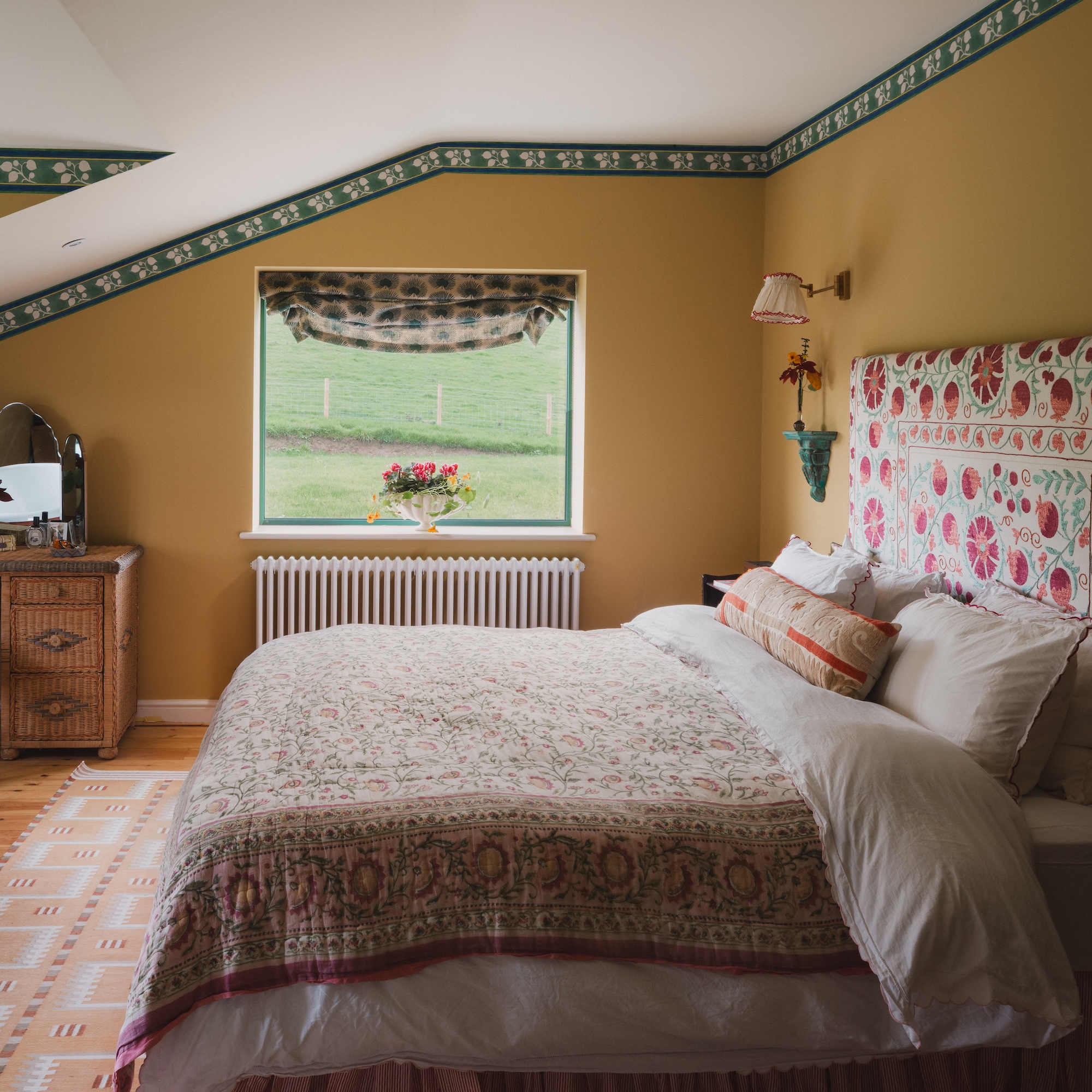yellow painted bedroom with green wallpaper border at top of wall, with upholstered bed and window overlooking grass