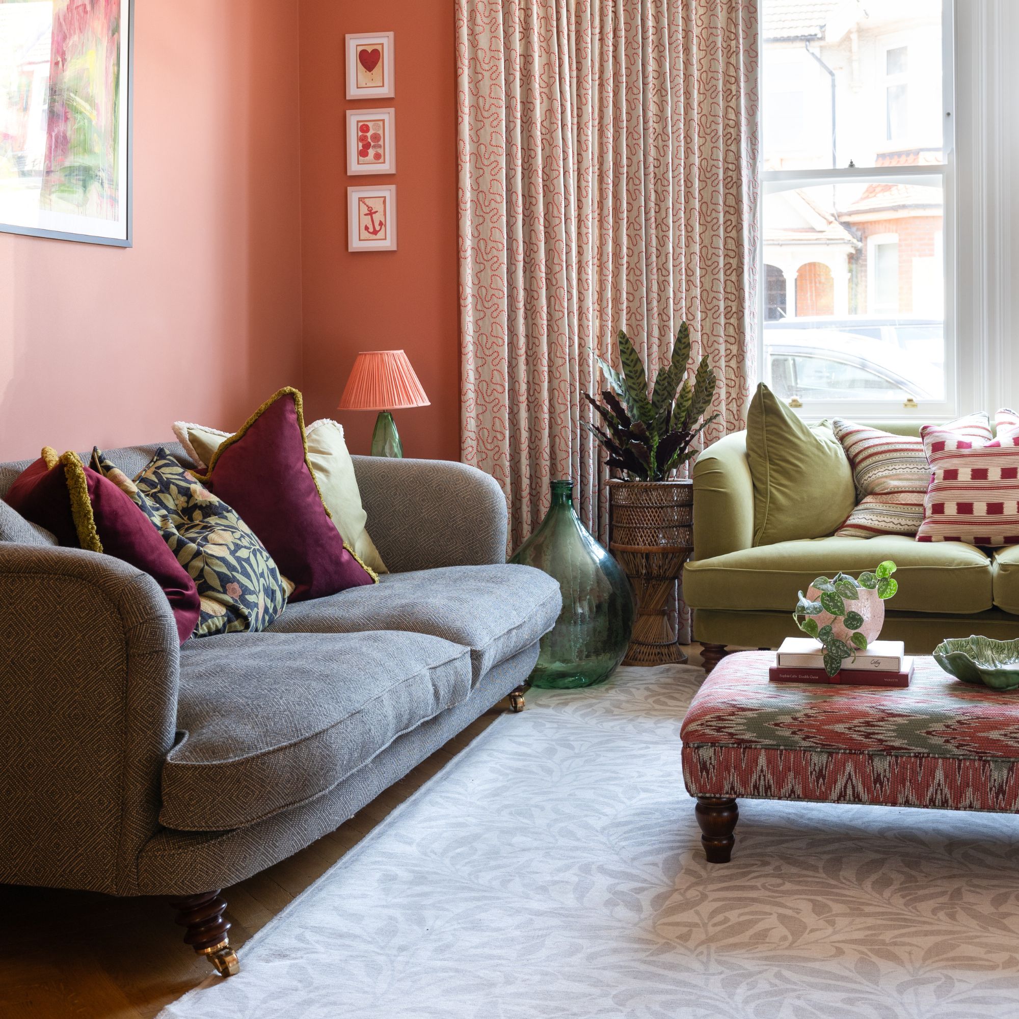 Pink period living room with a grey sofa next to a green sofa, and a patterned footstool, rug and curtains around the large window