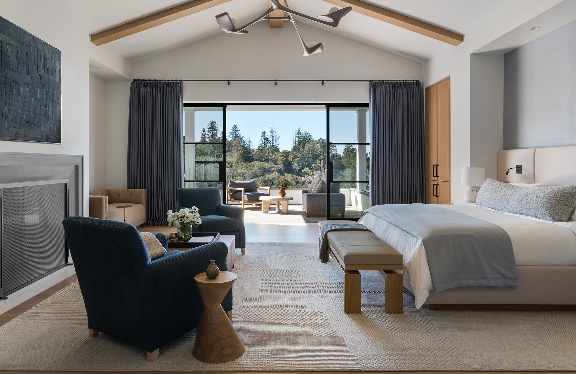 large modern bedroom with white bed and built-in lighting, bench seat at the foot of the bed, and two armchairs in front of fire, large rug, and pendant light, and large steel frame doors leading out to garden