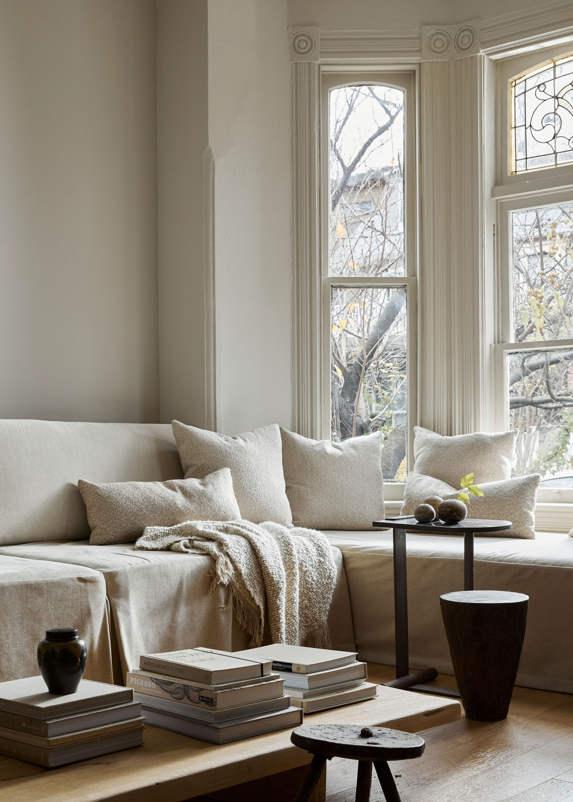 A cozy living room by bright windows with white soft pillows, a matching throw, a wood side table and stool, and a coffee table with book stacks