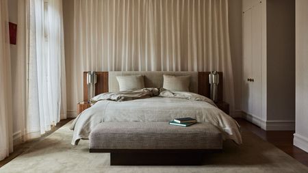 minimalist beige bedroom with curtains behind bed, ottoman at the end of the bed, rug, and timber extended headboard