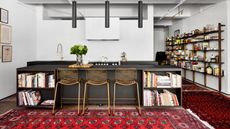 modern kitchen with black kitchen island, white walls, and brass bar stools, with red Persian rugs layered around the kitchen, with exposed bookshelves covered in books