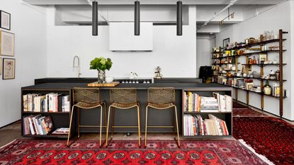 modern kitchen with black kitchen island, white walls, and brass bar stools, with red Persian rugs layered around the kitchen, with exposed bookshelves covered in books