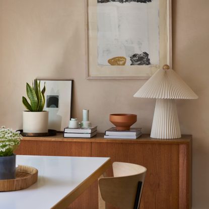 A wooden sideboard against a limewash wall with a pleated paper table lamp on top