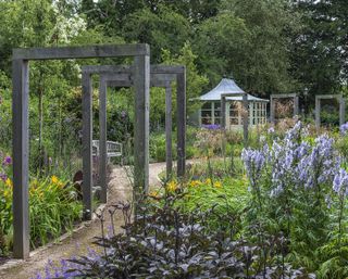 In the contemporary lower garden, a sinuous path of compacted aggregate leads beneath a pergola made up of eight standalone oak frames that, from either direction, frame views over the borders of aconites, dahlilies, dahlias, geums, sea hollies, catmint, campanulas, salvias, golden oats and verbenas