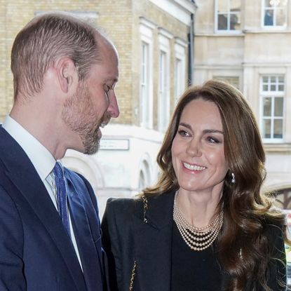 Kate Middleton has long, wavy brunette hair and wears a black jacket with a black top and a pearl necklace while gazing at husband Prince William's beard