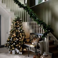Green hallway with faux fir garland wrapped around stair bannisters, faux fir Christmas tree light with fairy lights, silver baubles, presents, vintage style suitcases, wooden bench, wooden flooring, grey rug