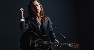 Jake Kiszka wears a dark suit, no shirt, and is photographed with his custom made Martin acoustic.